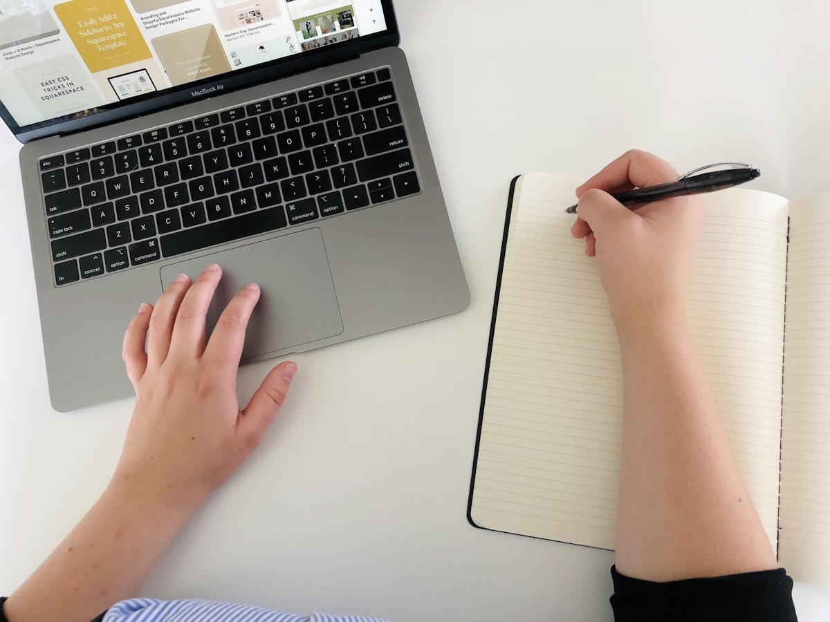 Person writing in a notebook while working on a laptop